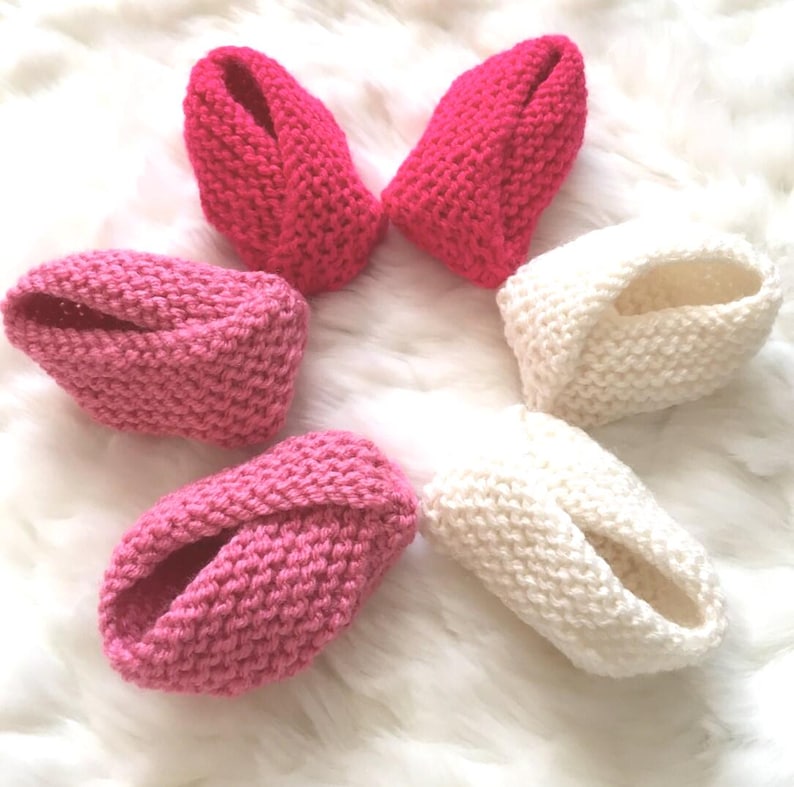 Puede incluir: Botines de beb&eacute; tejidos a mano en tonos rosa y blanco. Los botines est&aacute;n dispuestos sobre una superficie suave y blanca. La textura tejida es claramente visible, mostrando la artesan&iacute;a de los art&iacute;culos hechos a mano. Los botines est&aacute;n dise&ntilde;ados para mantener los pies del beb&eacute; calientes.