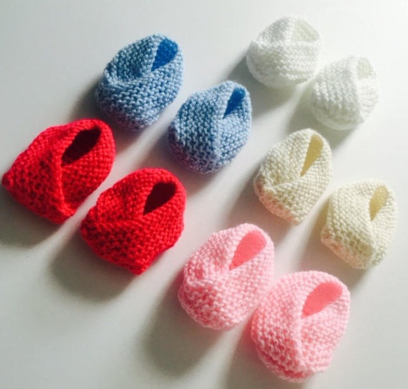 Puede incluir: Varios patucos de beb&eacute; tejidos a mano en rojo, azul claro, blanco, crema y rosa. El tejido suave y texturizado sugiere un dise&ntilde;o acogedor y c&oacute;modo para beb&eacute;s.