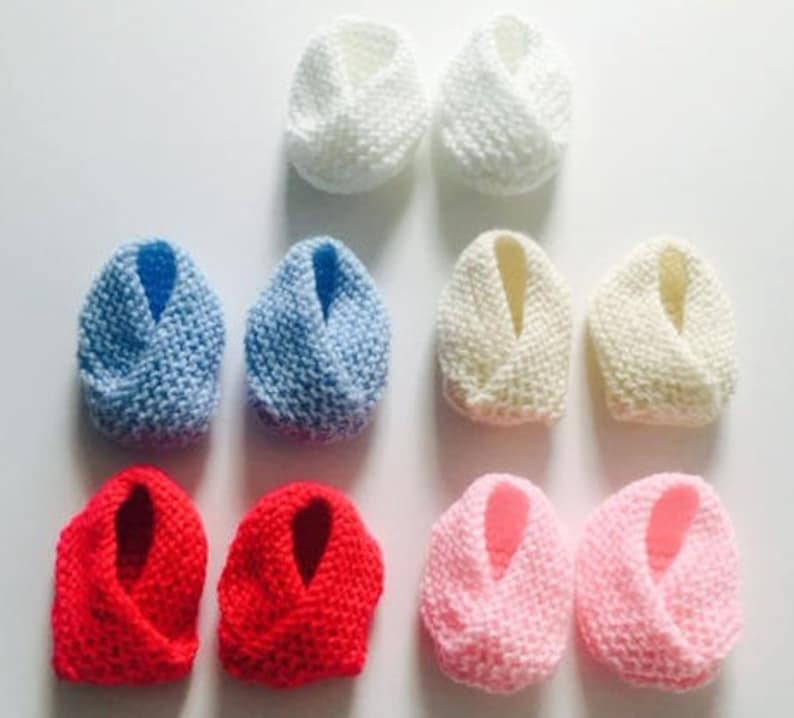 Puede incluir: Ocho pares de patucos de beb&eacute; de punto en varios colores. Los patucos est&aacute;n dispuestos en pares, mostrando tonos blancos, azules, crema, rojos y rosas. El tejido de punto suave y texturizado sugiere una sensaci&oacute;n acogedora y hecha a mano, ideal para reci&eacute;n nacidos.
