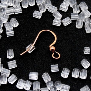 May include: A close-up of a gold earring hook with a clear plastic stopper. The earring hook is lying on a black surface with many clear plastic stoppers scattered around it.