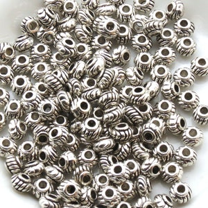 Op de afbeelding: Een grote stapel zilveren metalen kralen met een decoratief swirl-ontwerp. De kralen hebben een gat in het midden om te rijgen.