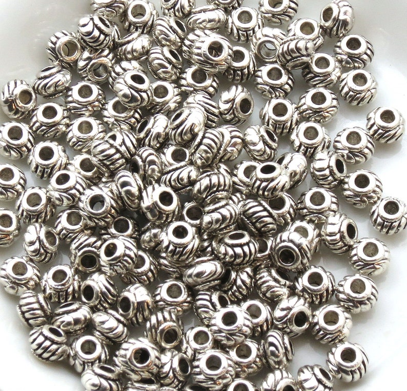 Op de afbeelding: Een grote stapel zilveren metalen kralen met een decoratief swirl-ontwerp. De kralen hebben een gat in het midden om te rijgen.