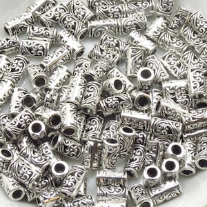 May include: A large pile of silver metal beads with an intricate, swirling design. The beads are cylindrical and have a hole through the center.
