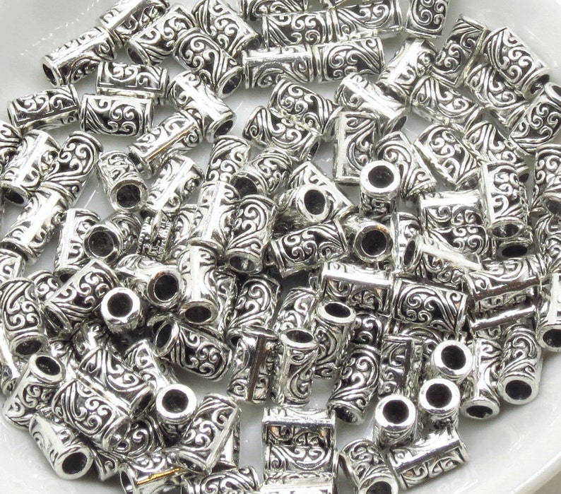 May include: A large pile of silver metal beads with an intricate, swirling design. The beads are cylindrical and have a hole through the center.