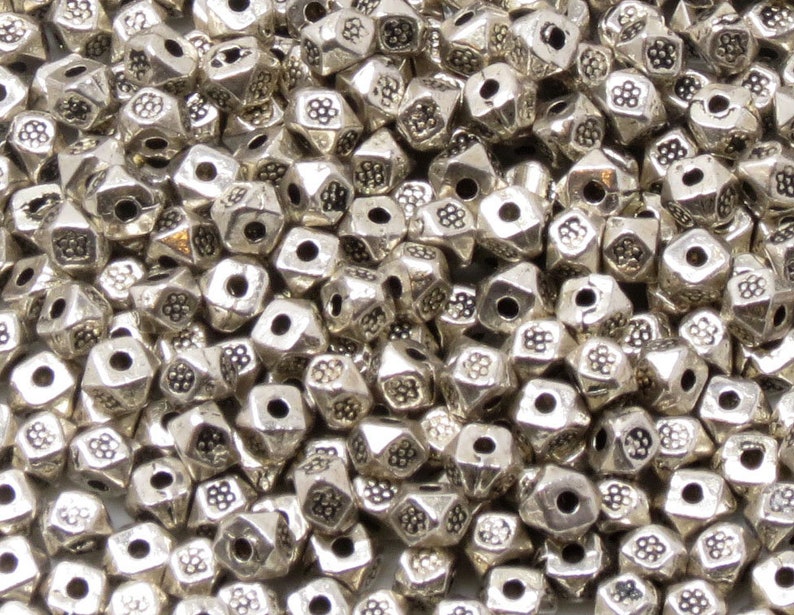 May include: A close-up of a pile of silver metal beads. The beads are shaped like a pyramid with a hole in the center. Each bead has a textured surface with a flower-like design.