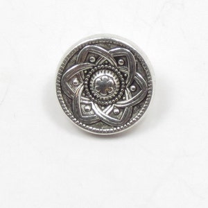 May include: A silver button with a detailed, intricate design featuring a central flower motif surrounded by a circular pattern.