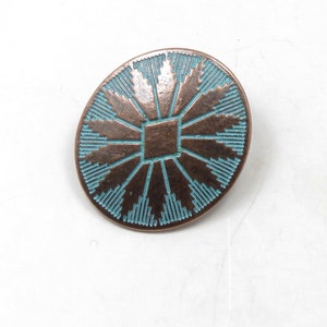 May include: A round copper-colored pin with a turquoise blue geometric design. The design features a central square with radiating lines.