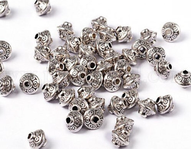 May include: A large group of silver metal beads with intricate designs. The beads are all the same size and shape, and they have a shiny finish.