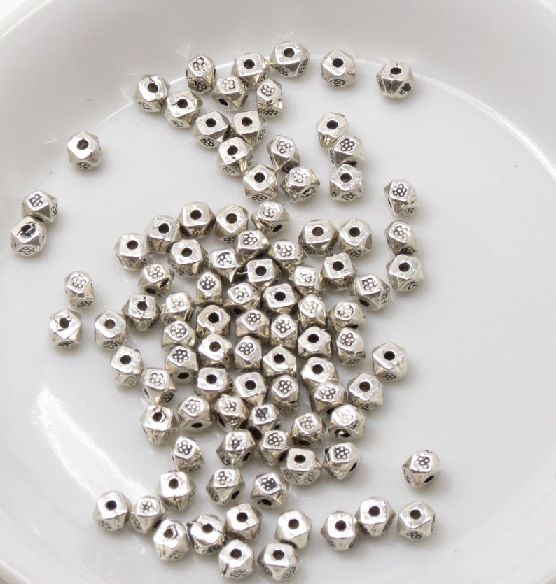 May include: A pile of silver metal beads with a flower design on each bead. The beads are scattered on a white surface.