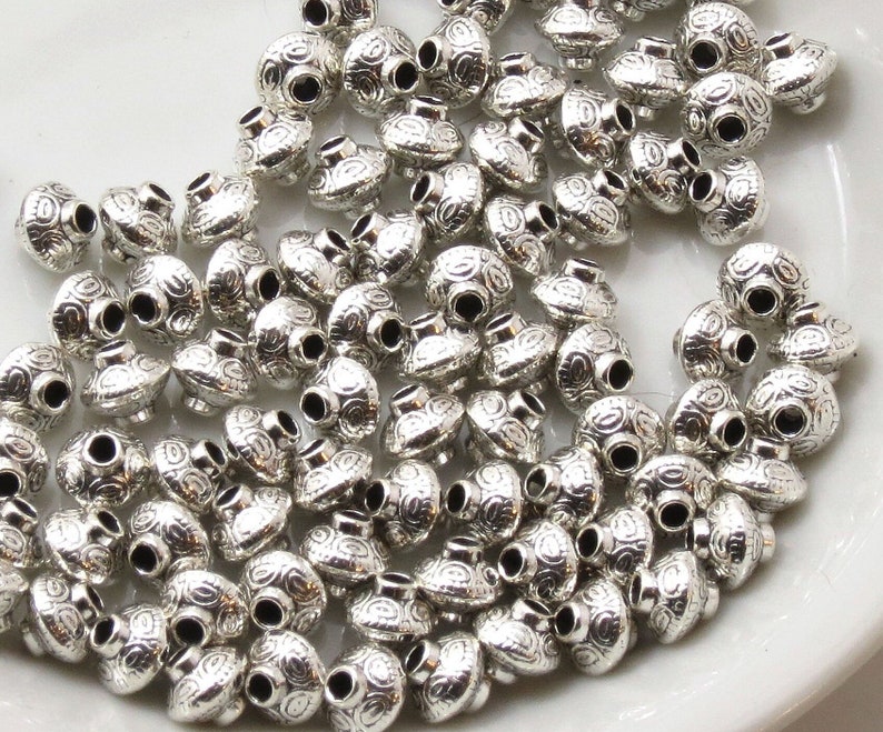 May include: A large quantity of silver metal beads with a decorative swirl pattern. The beads are round and have a hole in the center for stringing.