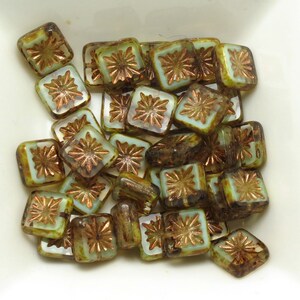 May include: A collection of square beads with a floral design. The beads have a light green and brown color scheme with a copper-colored flower in the center. The beads are in a white square dish.