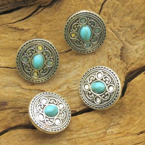 May include: Four silver-toned buttons with turquoise-colored oval center stones. The buttons have an ornate, circular design with a decorative border. The buttons are displayed on a wooden surface.