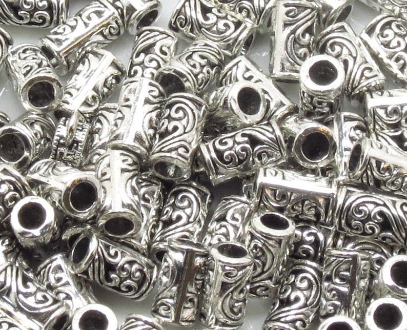May include: A pile of silver metal bead caps with an ornate design. The caps have a hole in the center and are designed to be used as end caps for jewelry making.