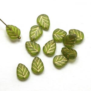 May include: A collection of green leaf-shaped beads with gold-colored vein detailing. One bead is attached to a wire. The beads are scattered on a white surface. These beads are suitable for jewelry making and other craft projects.