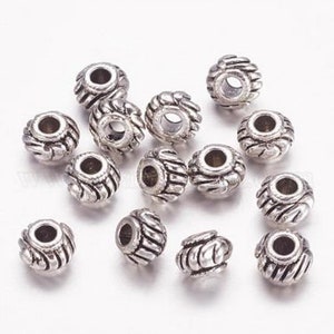 Op de afbeelding: Een groep van 15 zilveren metalen kralen met een decoratief ontwerp. De kralen zijn rond en hebben een gat in het midden om te rijgen.
