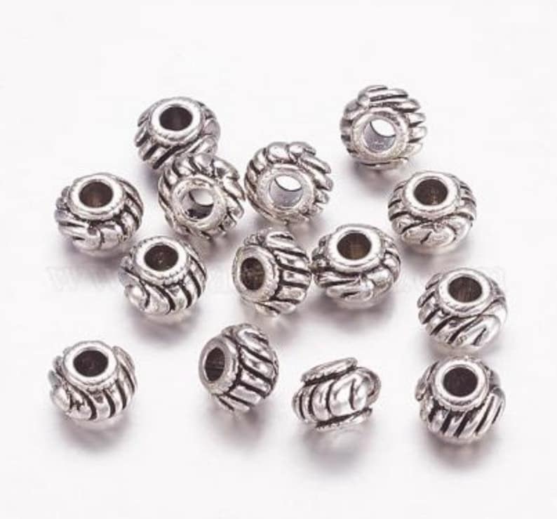Op de afbeelding: Een groep van 15 zilveren metalen kralen met een decoratief ontwerp. De kralen zijn rond en hebben een gat in het midden om te rijgen.