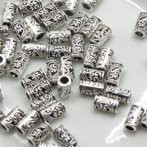 May include: A pile of silver-toned metal beads with a decorative swirl design. The beads are cylindrical and have a hole in the center for stringing.