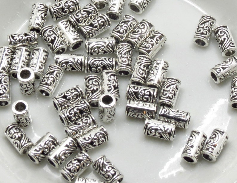 May include: A pile of silver-toned metal beads with a decorative swirl design. The beads are cylindrical and have a hole in the center for stringing.