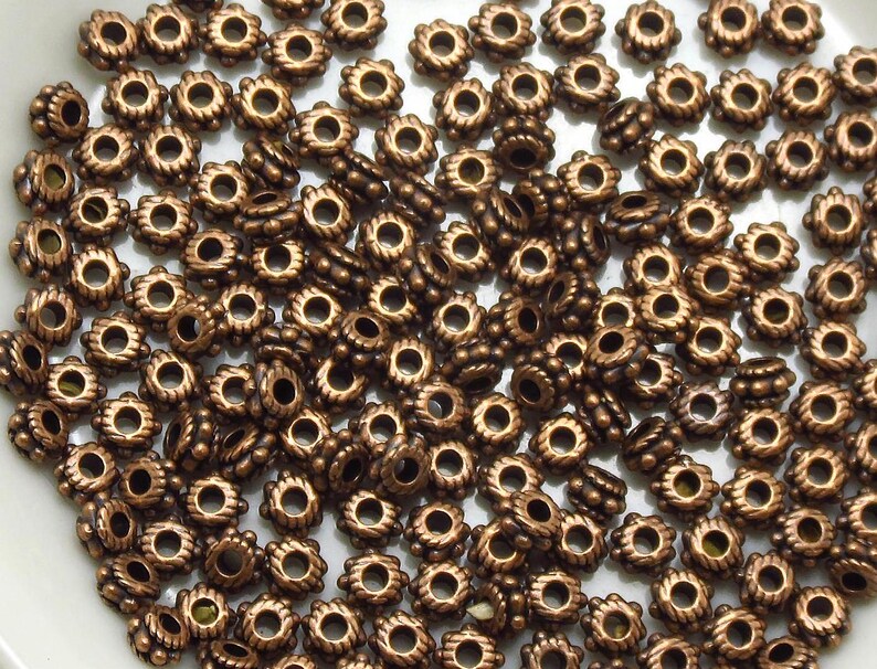 Puede incluir: Una colecci&oacute;n de cuentas de metal de color bronce. Las cuentas tienen un dise&ntilde;o floral con un agujero central y un borde texturizado en forma de cuerda. Algunas cuentas son cil&iacute;ndricas con una serie de cuentas redondas m&aacute;s peque&ntilde;as.