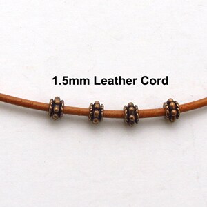Puede incluir: Primer plano de un cord&oacute;n de cuero marr&oacute;n claro con cuatro cuentas de bronce antiguo. El texto "1.5mm Leather Cord" est&aacute; impreso en negro. Las cuentas tienen un dise&ntilde;o detallado y texturizado.