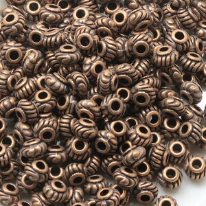 May include: A large pile of antique copper-colored metal beads with a decorative design. The beads have a hole in the center for stringing.