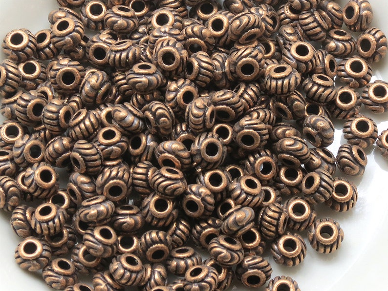 May include: A large pile of antique copper-colored metal beads with a decorative design. The beads have a hole in the center for stringing.
