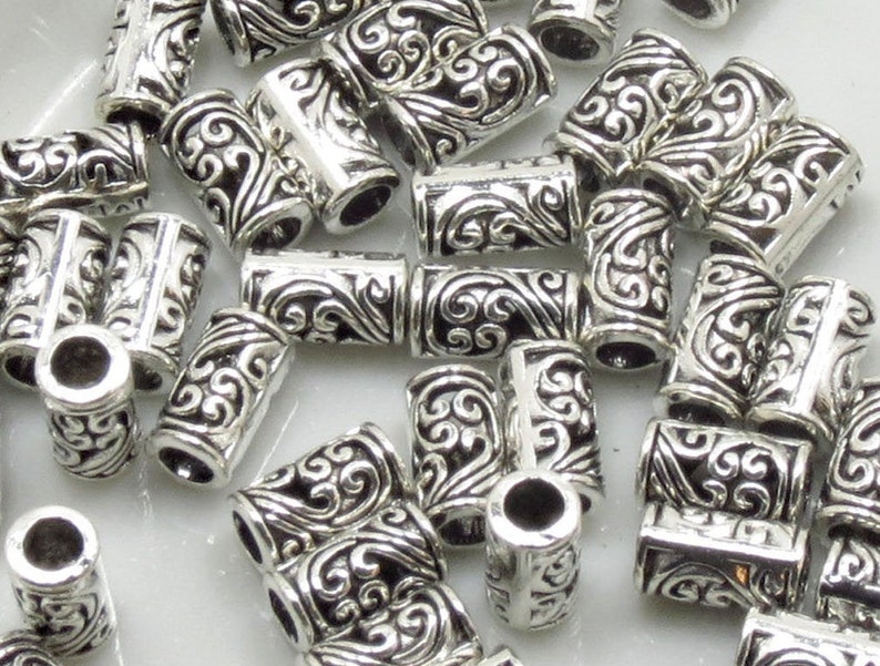 May include: A pile of silver-toned metal beads with a decorative, swirling design. The beads are cylindrical and have a hole through the center.