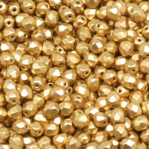 May include: Close-up of a pile of gold-colored faceted beads. The beads have a metallic finish and a geometric, multi-sided shape. These beads are suitable for jewelry making and craft projects.