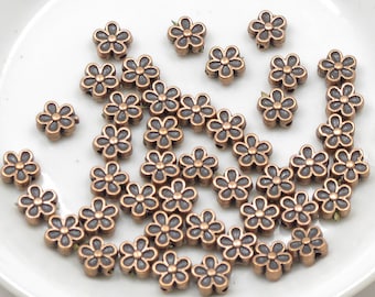 May include: A collection of copper-toned metal flower-shaped beads. The beads have a dark gray center and are scattered on a white surface.