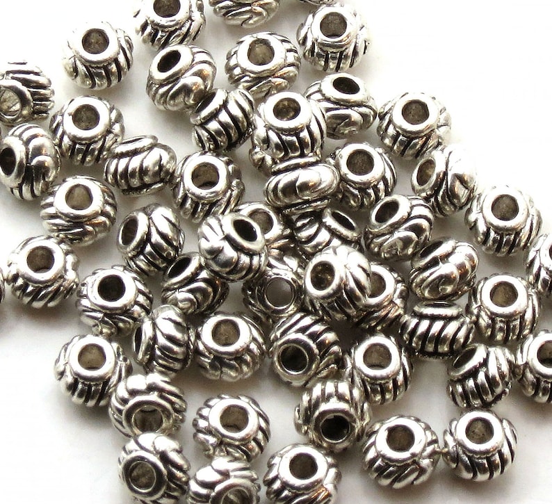 Op de afbeelding: Een stapel zilveren metalen kralen met een decoratief ontwerp. De kralen zijn rond met een gat in het midden.