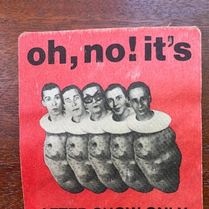 Op de afbeelding: Een rode stoffen patch met de tekst "oh, no! it's AFTER SHOW ONLY DEVO" in witte en zwarte letters. Vijf mannengezichten zijn zichtbaar in het midden van de patch.