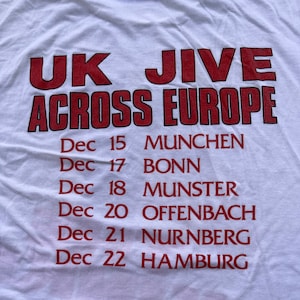 Könnte beinhalten: Ein weißes T-Shirt mit rotem Text, der "UK JIVE ACROSS EUROPE" lautet und Tourdaten im Dezember für Städte in Deutschland auflistet: München, Bonn, Münster, Offenbach, Nürnberg und Hamburg.
