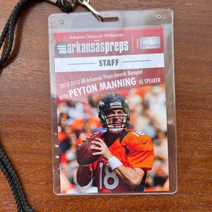 May include: A white plastic lanyard with a black cord and a clear plastic badge holder. The badge holder has a red and white logo that says "arkansaspreps" and a photo of Peyton Manning in a Denver Broncos uniform. The text on the badge reads "2012-2013 All Arkansas Preps Awards Banquet with Peyton Manning as Speaker".