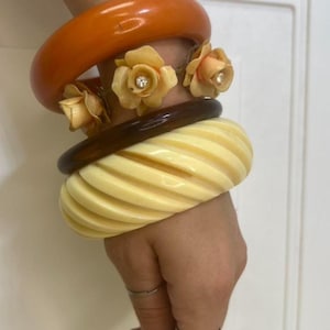 May include: A stack of four bangles including a thick orange band, a thin dark brown band, and a wide cream-colored band with a diagonal ribbed texture. Two pale peach-colored floral accents with small clear rhinestones are attached to the orange band.