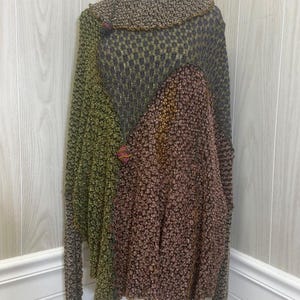 Puede incluir: Un poncho de punto de manga larga con un diseño de bloques de color y texturas múltiples en tonos verde oliva, gris carbón y marrón cobre intenso. La prenda incluye un cuello ancho doblado y pequeños detalles decorativos de punto colgantes.