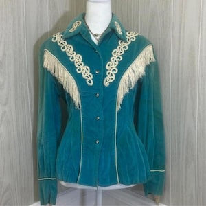 May include: A teal long-sleeve button-down shirt featuring cream-colored decorative embroidery and fringe detailing along the chest and shoulders. The shirt has a pointed collar, long sleeves with cuffs, and a structured fit with vertical seams.