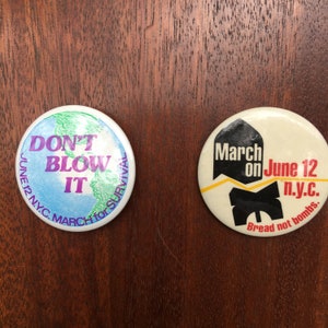 May include: Two round button pins with political messages. The left pin is blue, green, and purple with the text "Don't Blow It" and "June 12 N.Y.C. March for Survival". The right pin is white with black text that reads "March on June 12 n.y.c. Bread not bombs."