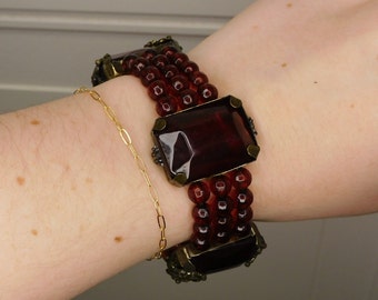 May include: A multi-strand bracelet with dark red glass beads and large, rectangular, dark red glass stones set in gold-toned metal.