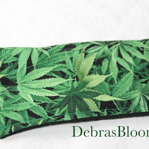 Può includere: Articolo rettangolare in tessuto con un motivo a foglie di cannabis verdi su sfondo nero. Ha un bordo nero e il testo "DebrasBlooms" in verde. Probabilmente si tratta di una custodia o di un accessorio simile.