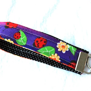 May include: A fabric wrist key fob with a purple background featuring red ladybirds, green leaves, and yellow flowers. The key fob has a black and white polka dot pattern on the underside and a silver metal clasp and key ring.