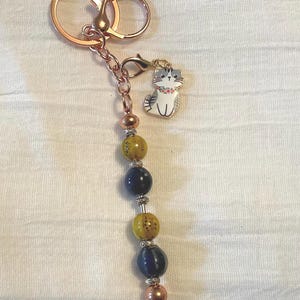 May include: A rose gold keychain with a cat charm and a beaded strand. The cat charm is white, gray, and black with a pink collar. The beaded strand has yellow, blue, and silver beads.