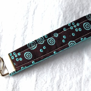 May include: A fabric key fob with a brown background and teal circular patterns. The key fob has a silver metal clasp and a key ring. The key fob is approximately 6 inches long.