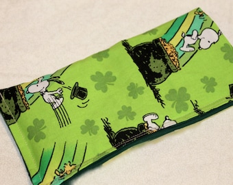 May include: A rectangular, green fabric item featuring a St. Patrick's Day theme. The design includes Snoopy characters, a pot of gold, shamrocks, and a leprechaun's hat. The item has a dark green border and is likely a decorative item.