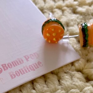 May include: A pair of orange and green hamburger earrings with a white post. The earrings are on a white card with the text "Bomb Pop Boutique" in pink.