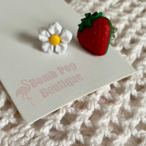 May include: Two small, colourful, plastic earrings. One earring is a white daisy with a yellow centre. The other earring is a red strawberry with green leaves. The earrings are on a white card with the text "Bomb Pop Boutique" and a pink flamingo.