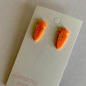 May include: A pair of orange carrot-shaped stud earrings. The earrings are on a white card with the text "Bomb Pop Boutique" printed on it.