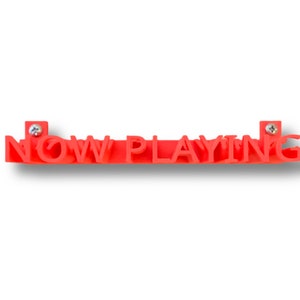 May include: A red plastic sign with the words "NOW PLAYING" in raised letters. The sign is attached to a wall with two screws.