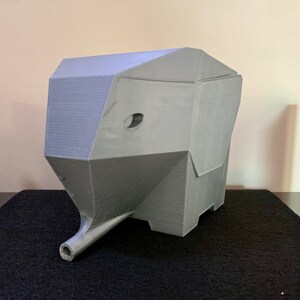 May include: A gray, geometric elephant head sculpture with a trunk and a small circular eye. The sculpture is 3D-printed and sits on a black surface. The elephant head is a unique piece of home decor.