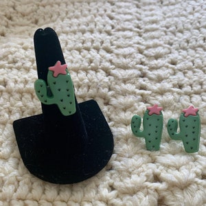 May include: Three green cactus earrings with pink star accents. The earrings are on a black display stand.