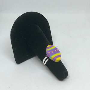 May include: A colorful Easter egg ring with a silver band. The egg-shaped top features a yellow base with purple and green stripes and dots. The ring is displayed on a black velvet ring holder.